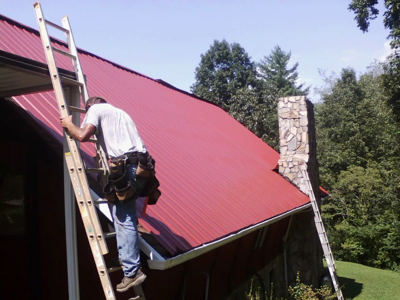 Roofer on a ladder beside a red metal roof and stone chimney — Soffit & Fascia Repair by Roofing Brothers in Ambler